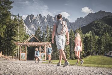 Due bambini giocano in un parco giochi davanti a un paesaggio montano; in primo piano una donna cammina su un sentiero di ghiaia, mentre sullo sfondo si vedono un prato, un bosco e alte cime rocciose.