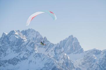 A paraglider is soaring above an expansive mountain landscape with snow-covered peaks.