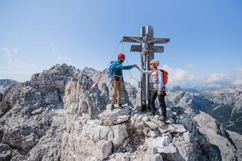 Two climbers are standing in front of a summit cross; in the background, mountain scenery can be seen, with the blue sky above.