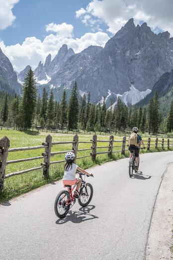 Una donna e un bambino viaggiano in bicicletta su una strada asfaltata attraverso una valle di montagna; sullo sfondo si vedono prati, una staccionata in legno, un bosco e alte cime montuose.