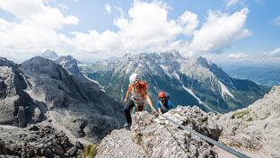 Due arrampicatori salgono su una roccia; sullo sfondo si vedono montagne, valli e il cielo azzurro con nuvole bianche.