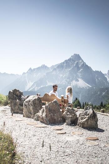 A man and a child are sitting on a wooden bench; an imposing mountain landscape can be seen in the background.