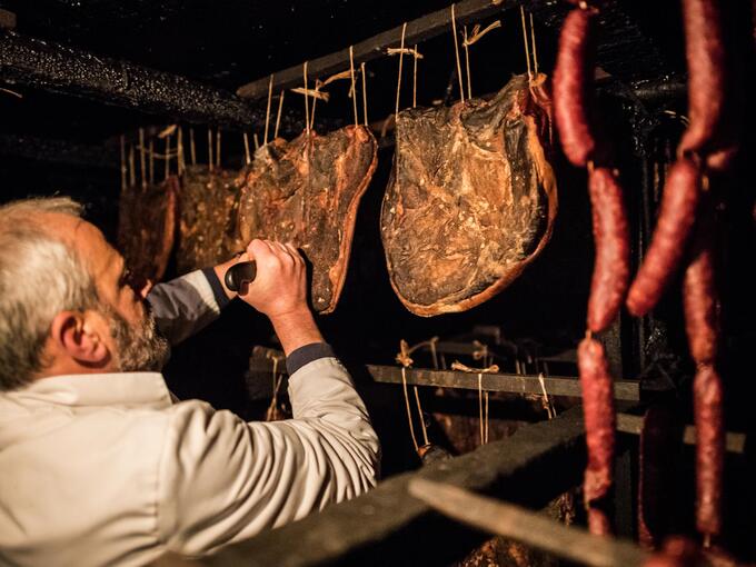 A man cuts hanging speck and cured sausages with a knife in a dark curing cellar