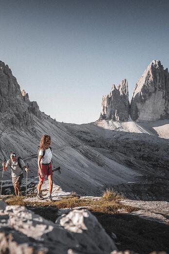 A man and a woman are hiking in the Dolomites, with the Three Peaks rising in the background