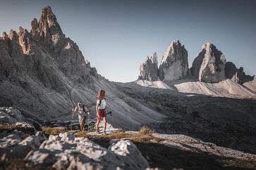 A man and a woman are hiking in the Dolomites, with the Three Peaks rising in the background