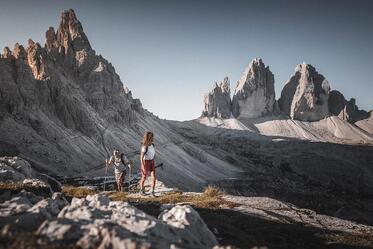 A man and a woman are hiking in the Dolomites, with the Three Peaks rising in the background