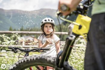Una bambina con il casco da bicicletta sta accanto a una bicicletta; in primo piano si vede sfocata un’altra bicicletta, sullo sfondo un muro di pietra e le montagne.