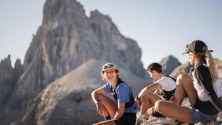 Three mountain runners are sitting in rocky terrain, looking out at the massive mountain landscape.