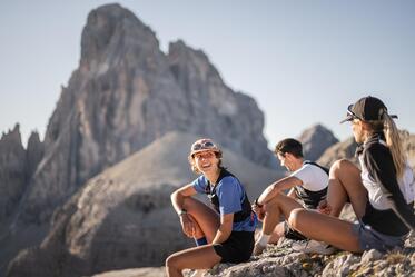 Tre trail runner siedono su un terreno roccioso e osservano l’imponente paesaggio montano.