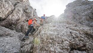 Three climbers are climbing a steep rock face; the sun and sky can be seen from above.
