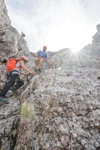 Tre arrampicatori scalano una ripida parete rocciosa; dall’alto si vedono il sole e il cielo.