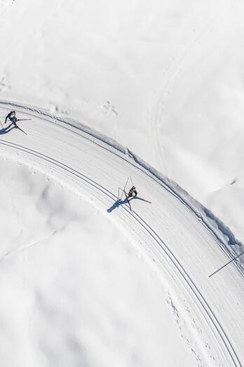 Ripresa aerea di una curva innevata con tracce nella neve e due persone su un percorso invernale.