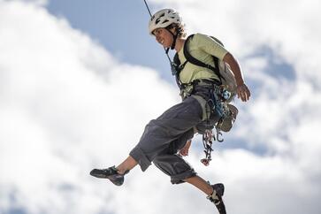 Climber with helmet and backpack hanging securely from a rope in front of a blue sky with white clouds.