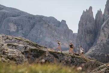 Three hikers in an expansive mountain range; a massive mountain landscape can be seen in the background.