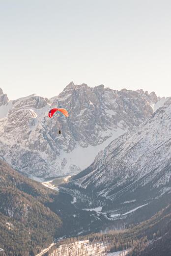 Two paragliders are soaring above an expansive mountain landscape with snow-covered peaks in the background.