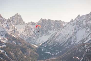 Two paragliders are soaring above an expansive mountain landscape with snow-covered peaks in the background.