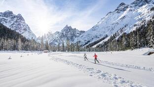Fondisti percorrono un’ampia pista da sci di fondo innevata in un soleggiato paesaggio invernale con montagne, bosco e cielo azzurro.