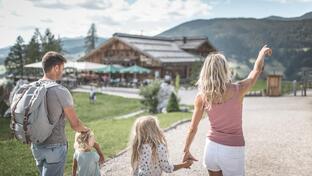 A family walks across a spacious square toward a building with a play area; trees, mountains and a cloudy sky can be seen in the background.