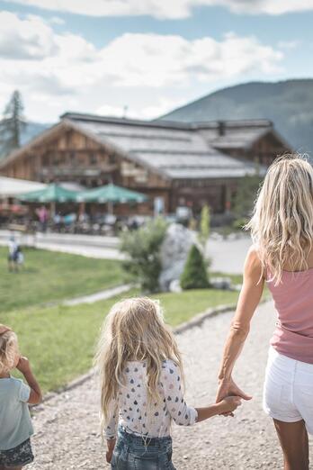 Una famiglia attraversa un ampio piazzale in direzione di un edificio con area giochi; sullo sfondo si vedono alberi, montagne e un cielo nuvoloso.