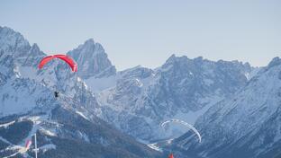 Two paragliders are flying in front of an expansive mountain landscape with snow-covered peaks.