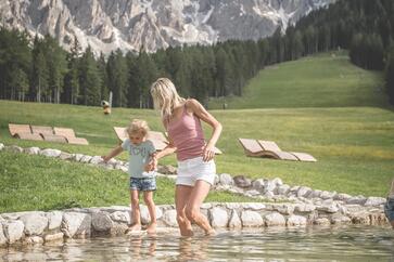 A woman and a child stand barefoot in a shallow stream in front of a mountain landscape; in the background, a meadow, a forest and striking rocky peaks can be seen.