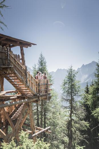 A man, a woman and two children sit on a wooden structure in the forest and look toward a mountain landscape.