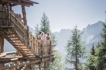 Un uomo, una donna e due bambini sono seduti su una struttura in legno nel bosco e guardano verso un paesaggio montano.