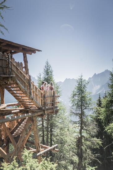 A man, a woman and two children sit on a wooden structure in the forest and look toward a mountain landscape.