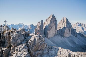Le Tre Cime con il paesaggio d’alta montagna circostante e il cielo azzurro sopra; a sinistra si vede una croce di vetta su un punto panoramico roccioso.