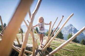 A child balances on a climbing frame made of wooden poles on a meadow; mountains, trees and a blue sky can be seen in the background.